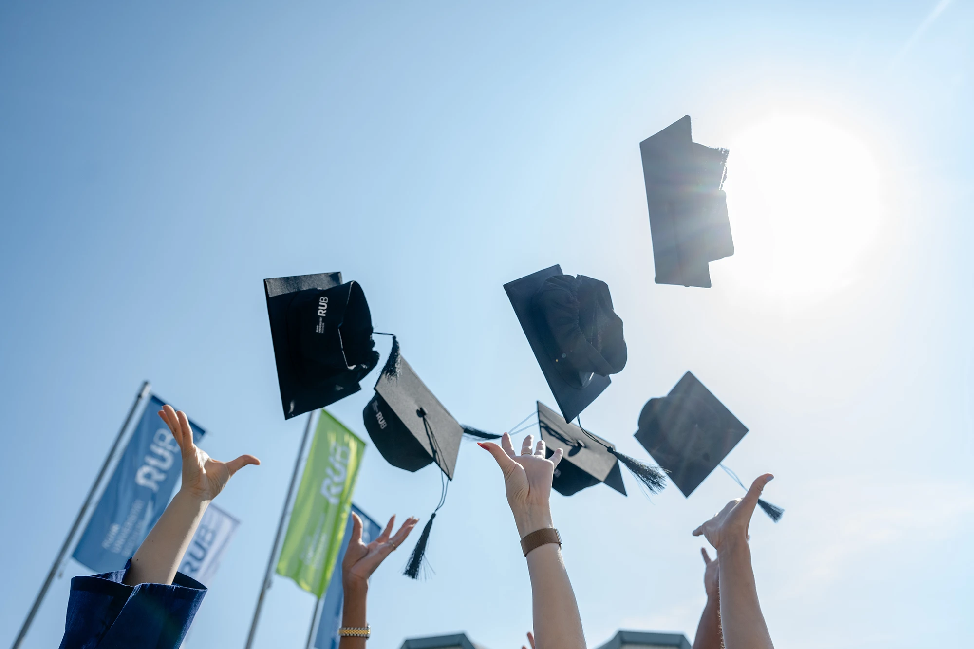 Graduates toss black mortarboards into blue sky during a sunny outdoor ceremony.
