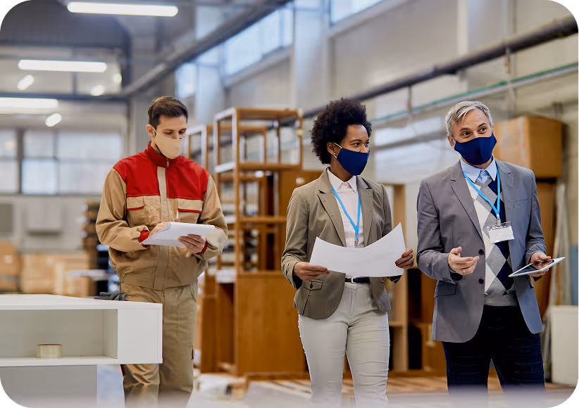 Three masked professionals in a warehouse reviewing documents and using a tablet.