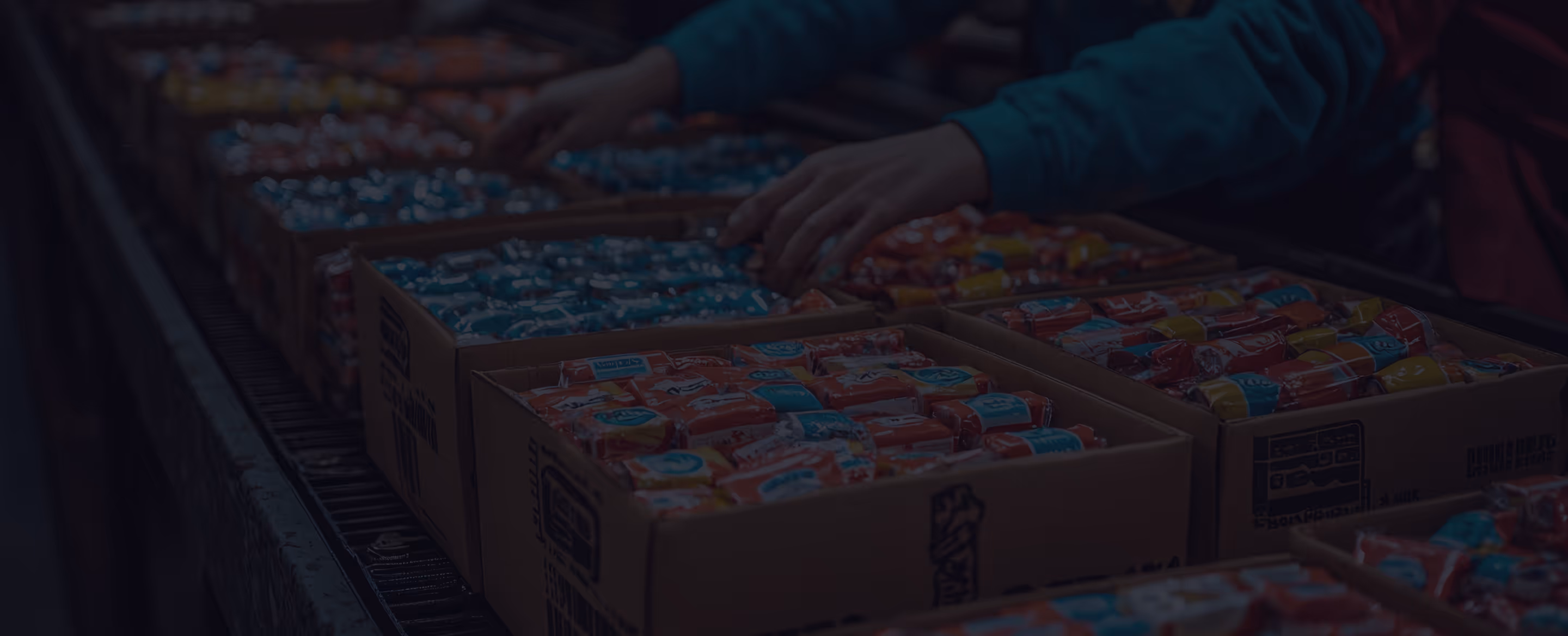 Hands organizing colorful packaged products in cardboard boxes on a conveyor belt in a factory.