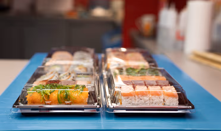 Two plastic trays of assorted sushi rolls on a blue surface with a blurred kitchen background.