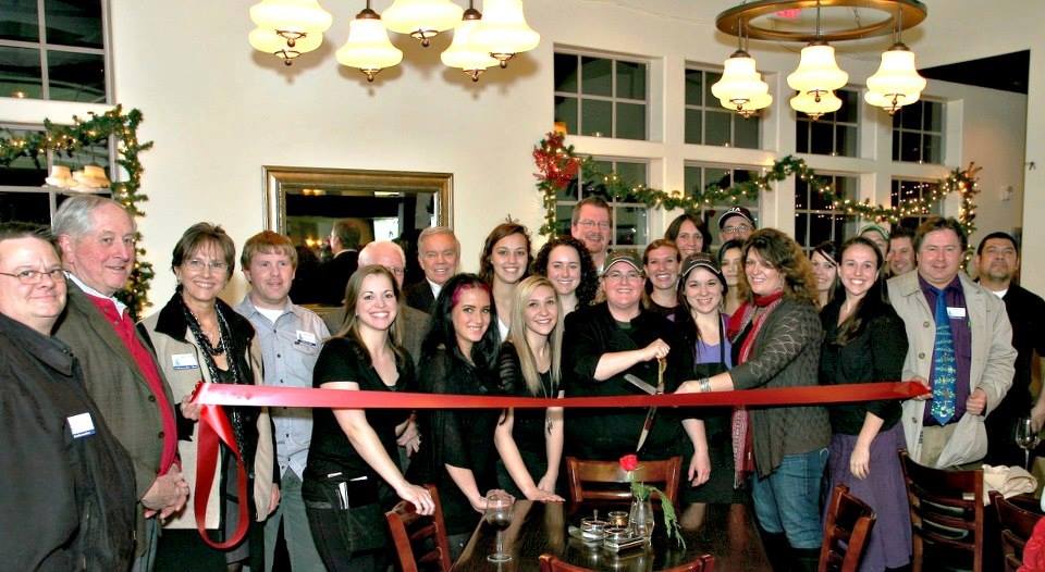 A large group stands behind a red ribbon at a ribbon cutting inside a restaurant decorated for the holidays.