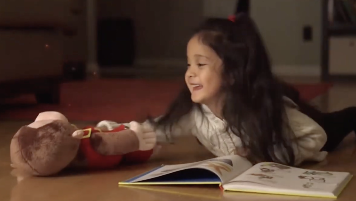 A young girl laughs on the floor beside a stuffed toy and an open picture book.