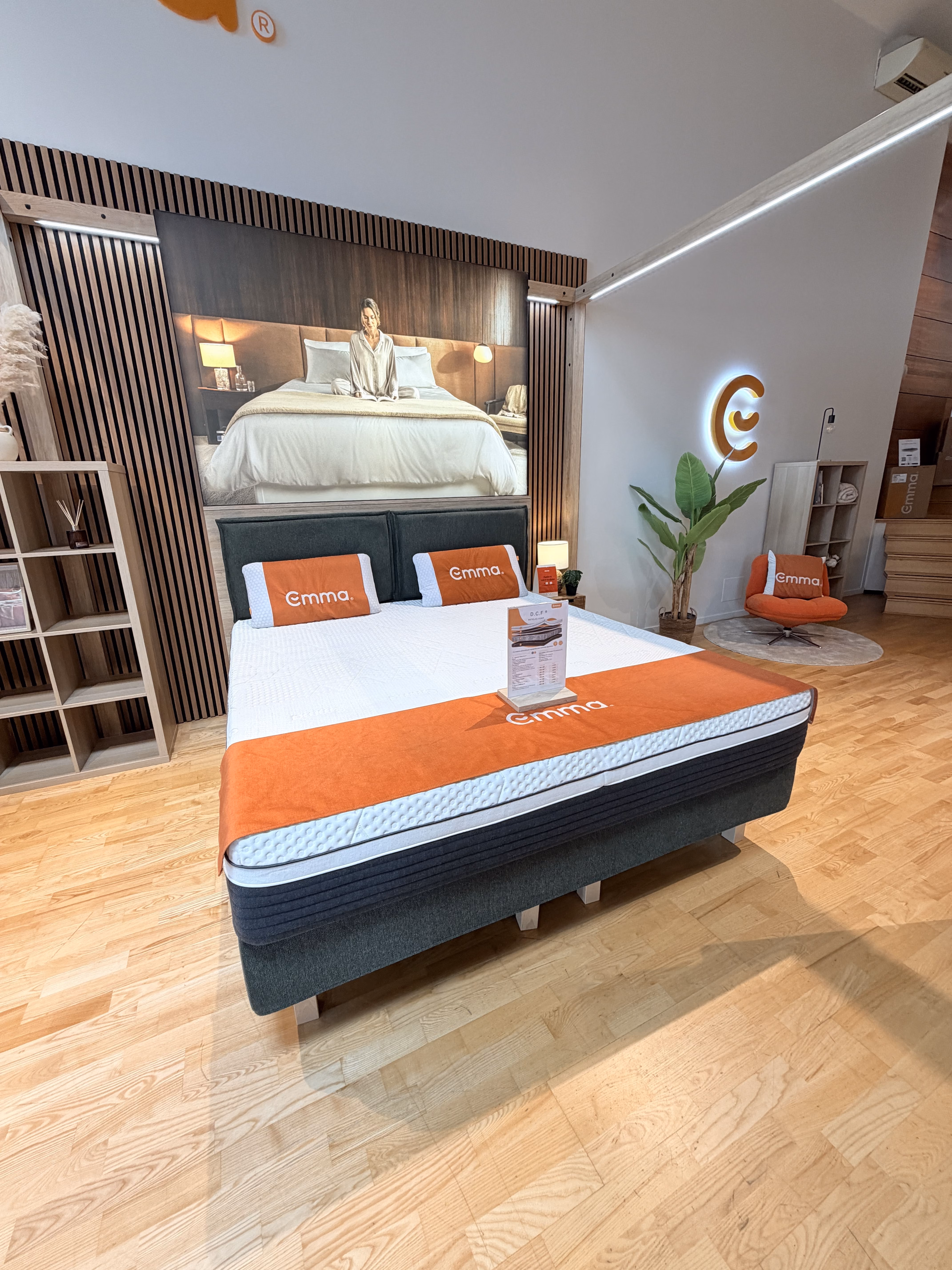 Modern bedroom showroom with Emma mattress, orange pillows, and a large framed photo on wooden floor.