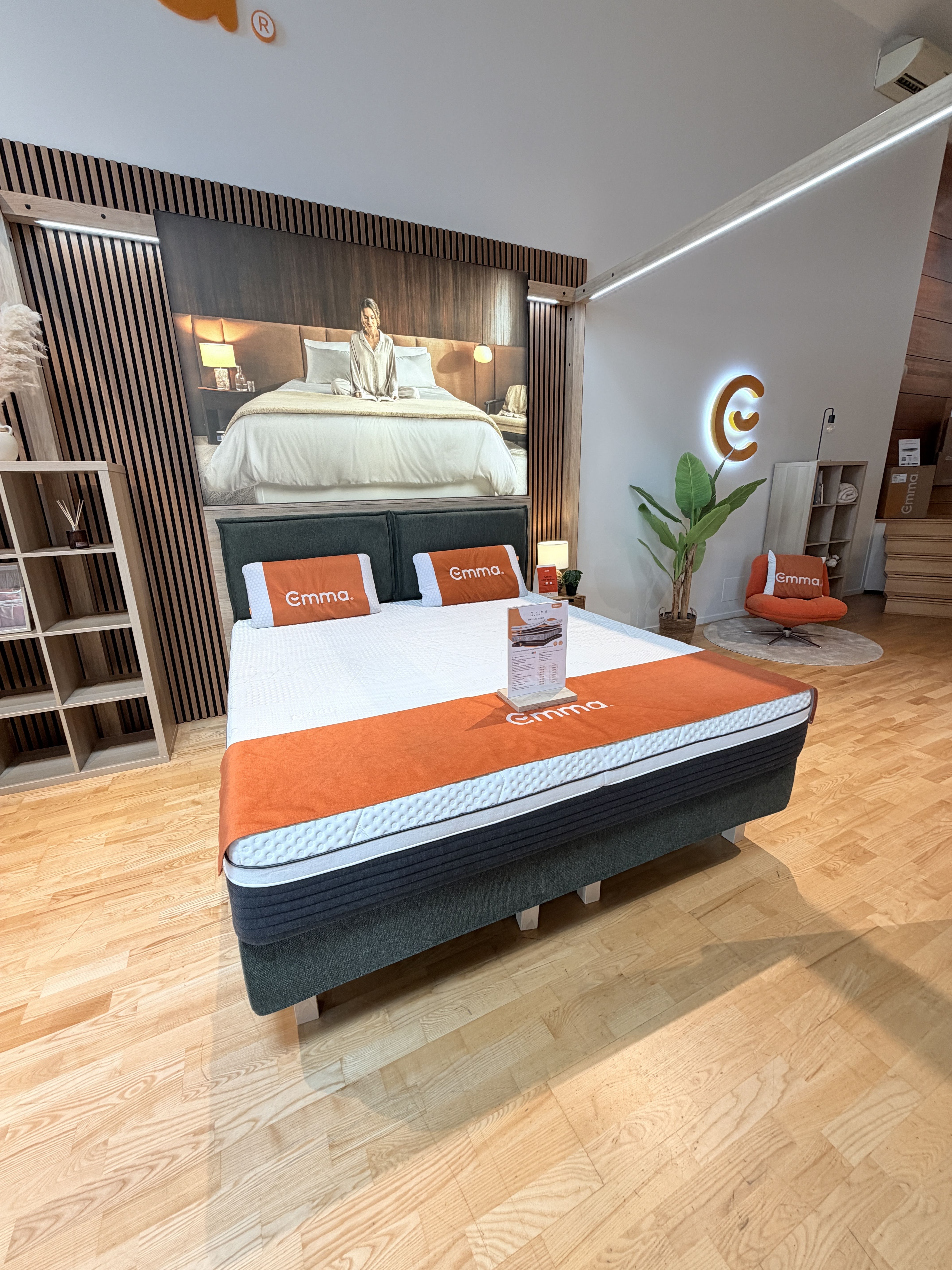 Modern bedroom showroom with Emma mattress, orange pillows, and a large framed photo on wooden floor.