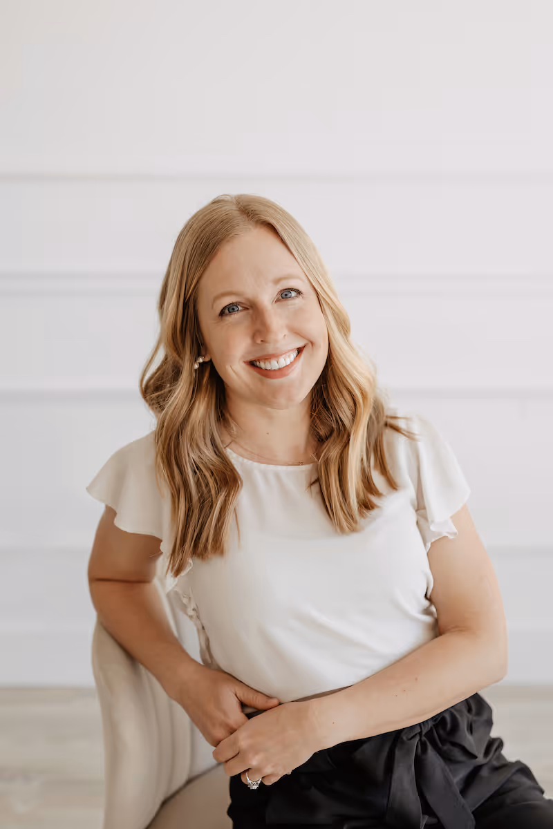 Smiling woman with wavy blonde hair wearing a white tshirt against a light background.