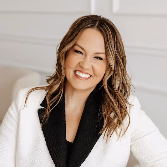 Smiling woman with wavy brown hair wearing a white jacket with black lapels against a light background.