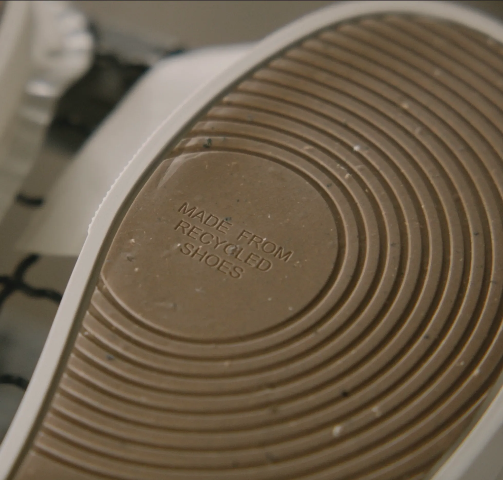 A detailed macro shot of the bottom of a shoe's sole. The beige rubber surface features concentric circular ridges for grip. In the centre, within a smooth circular section, the words "MADE FROM RECYCLED SHOES" are embossed in clean, capitalised sans-serif lettering. The material shows tiny dark flecks, indicating its recycled composition.