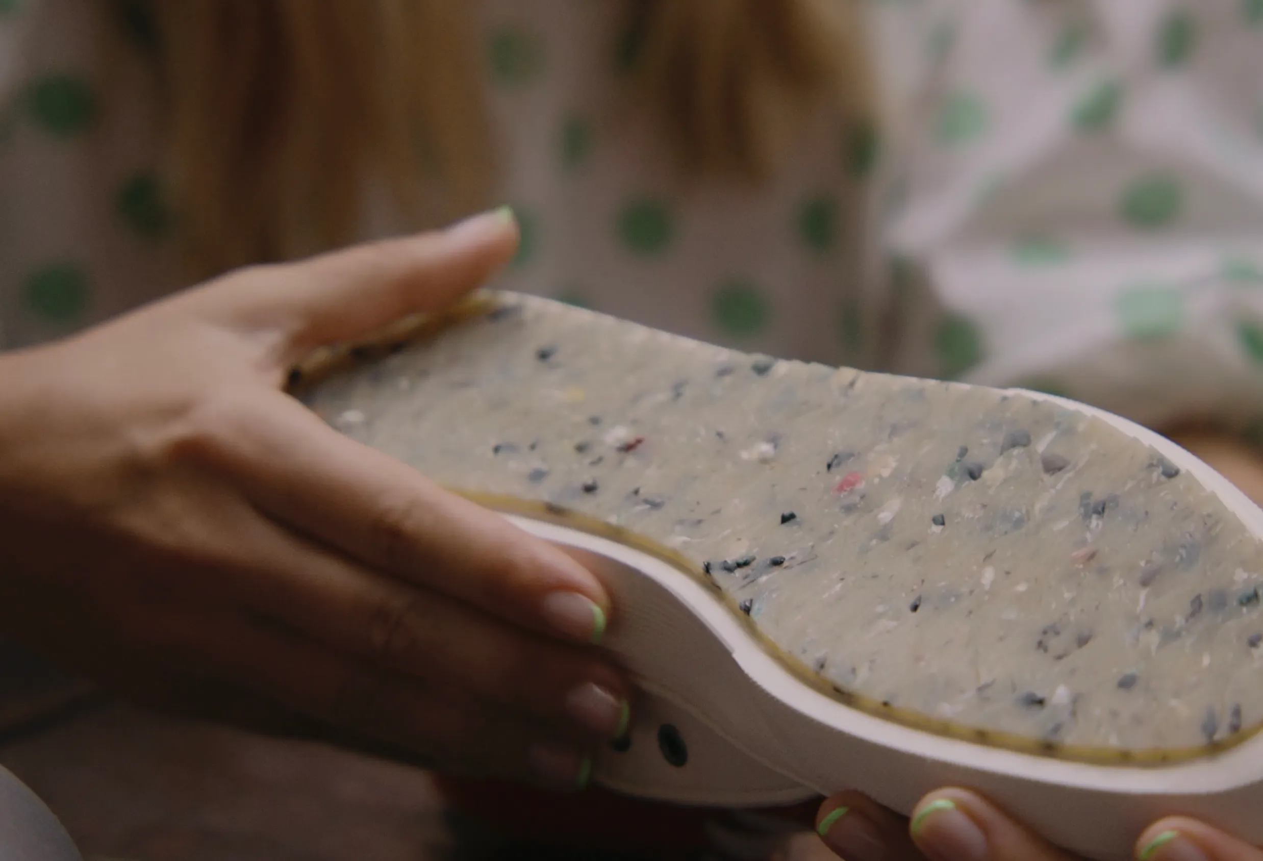A close-up shot of someone holding a shoe upside down to reveal its unique outsole. The sole has a translucent, yellowish-grey appearance with a multicoloured speckled texture, containing visible recycled fragments in shades of blue, black, and red. The person holding the shoe has neon green tipped nails, and a blurred background shows a person wearing a green polka-dot shirt.