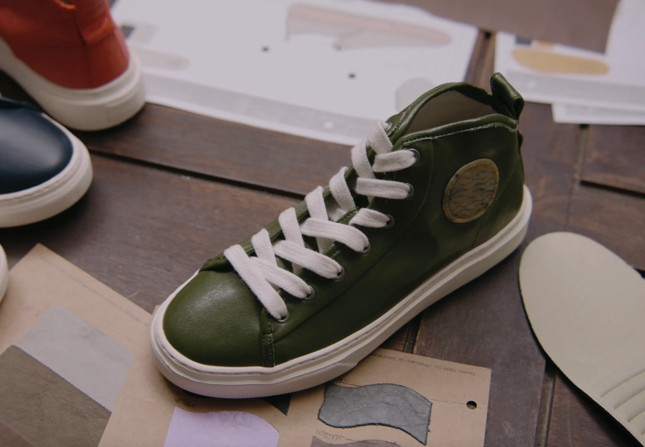 A top-down view of a dark wooden workshop table covered with shoe design materials. In the centre, an olive green leather high-top trainer with white laces sits on top of technical sketches and material swatches. Other trainers in navy blue and orange are partially visible in the corners, alongside a loose off-white insole. The scene suggests a creative design process in a studio.