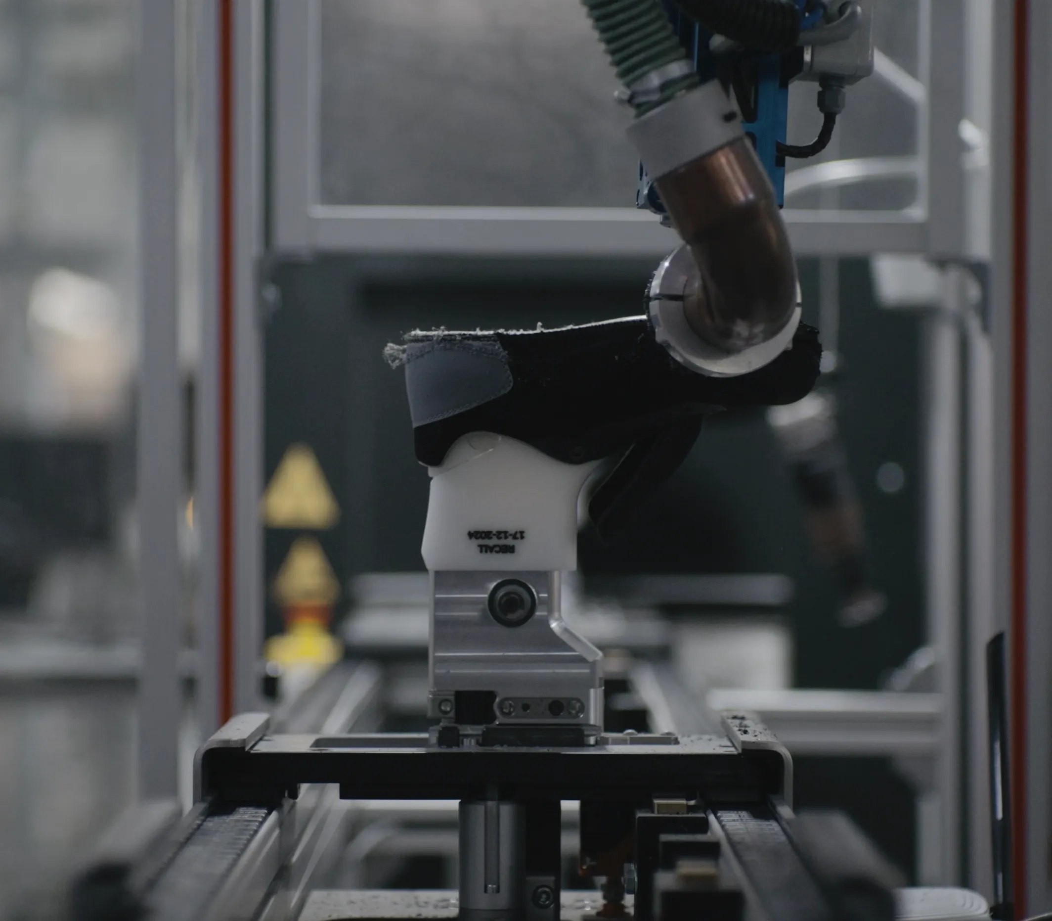 A clinical, industrial shot of a robotic assembly line in a factory. A mechanical arm with metal and white components holds a black shoe upper firmly in place over a conveyor system. The machinery is complex, featuring wires and metallic structures, with a soft-focus industrial background. The white part of the robotic grip has small black text and a date printed on it, reflecting a high-tech manufacturing environment.