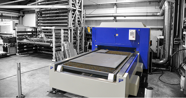 Industrial laser cutting machine in a factory with steel sheets on its bed and metal racks in the background.