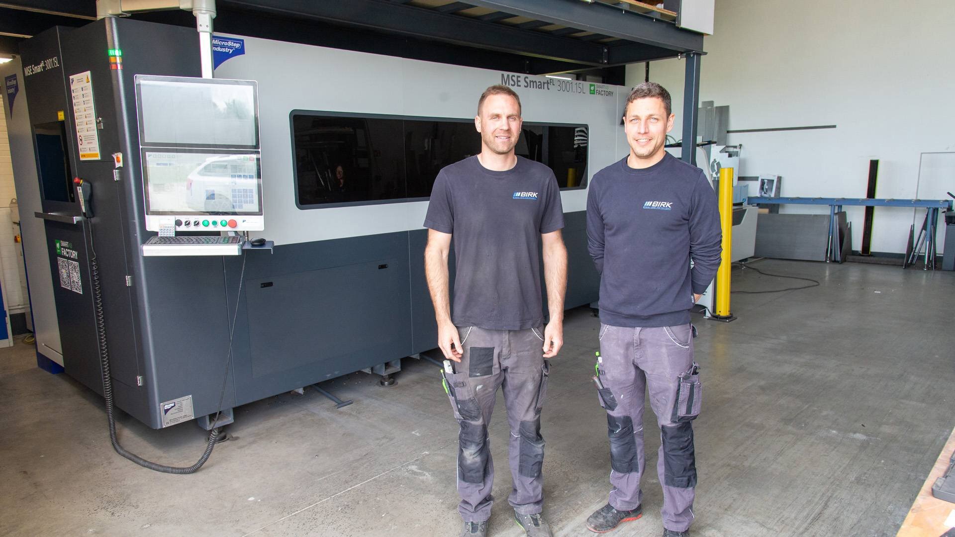 Two workers in black BIRK shirts standing inside a factory beside a large industrial machine.