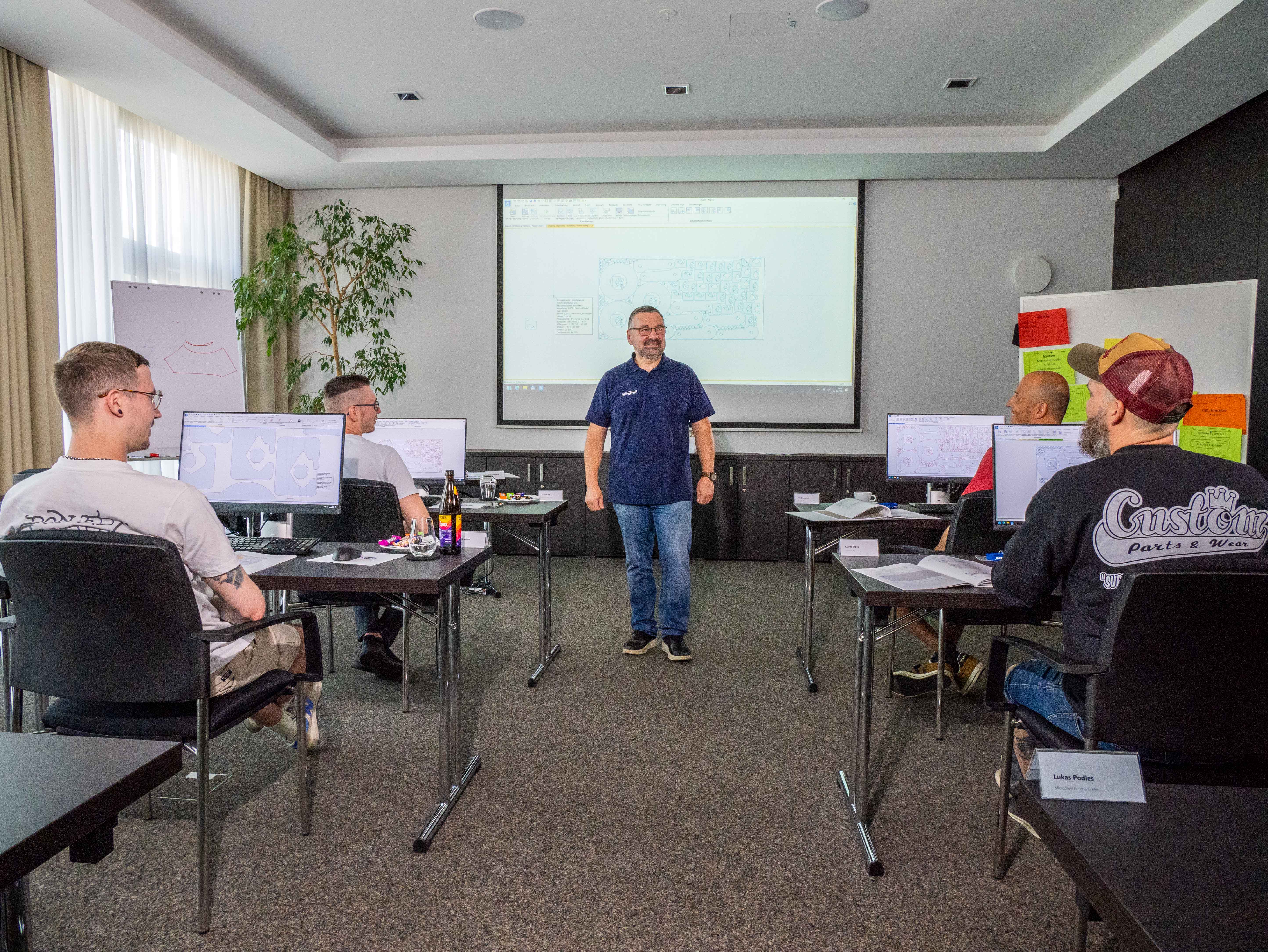 Man standing at front of classroom with seated students using computers and a projection screen.