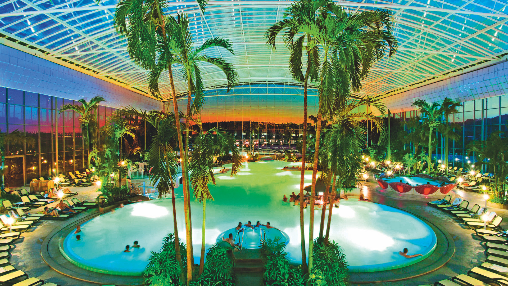 Indoor tropical-themed pool area with palm trees, lounge chairs, and people relaxing in pools at sunset.