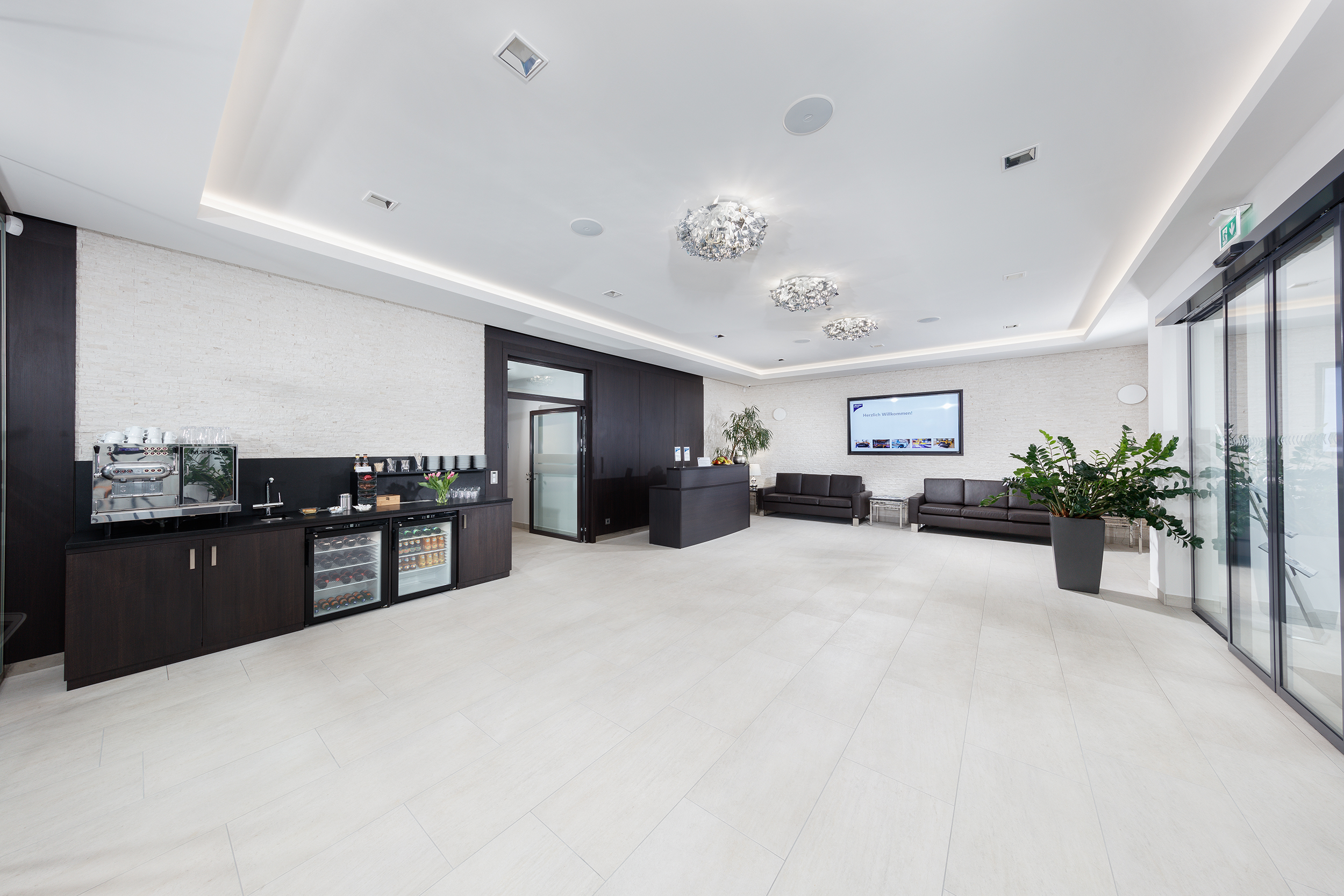 Modern office lobby with coffee station, reception desk, leather sofas, large plant, and glass entrance doors.