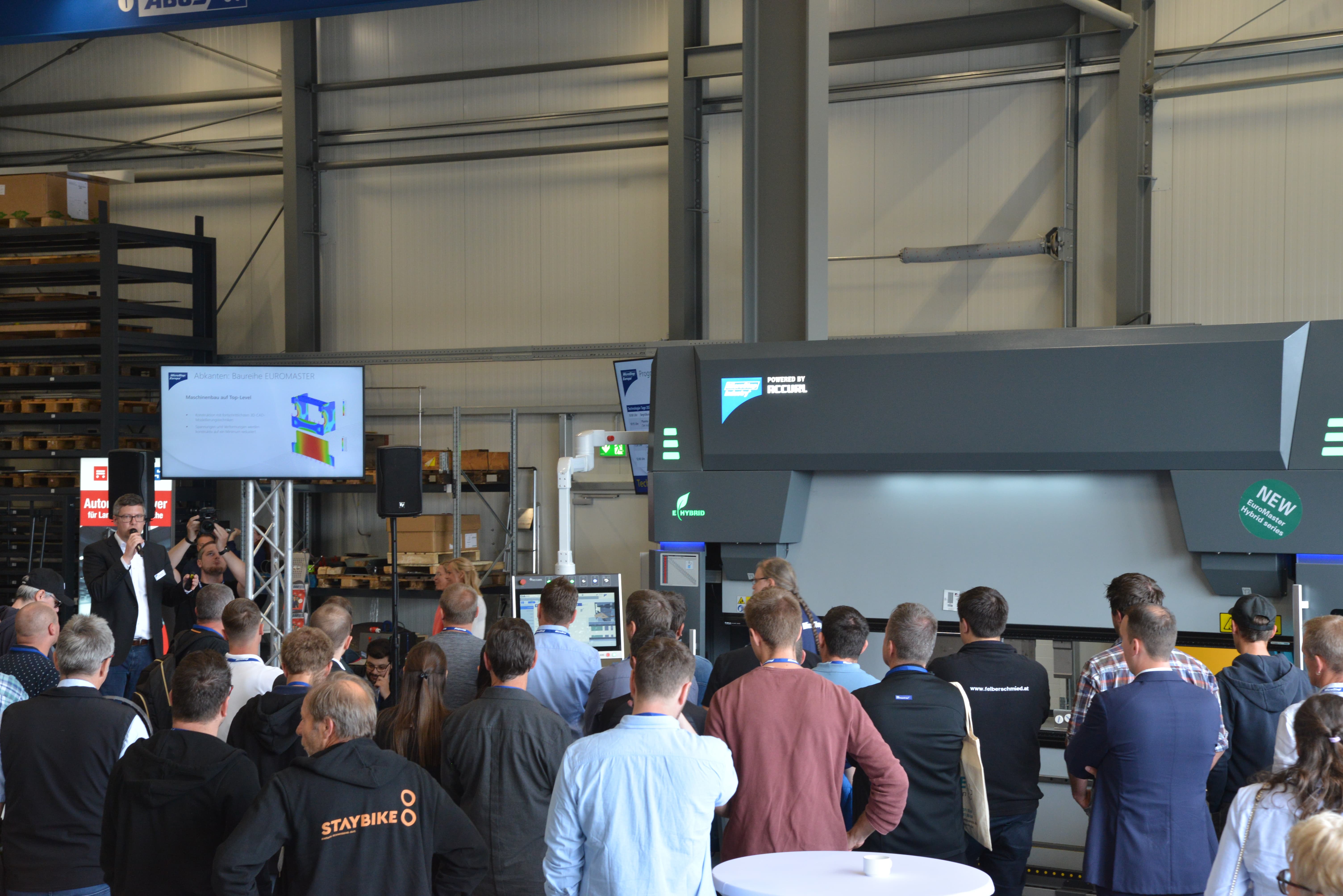 Audience watches a man speaking at an industrial machine demonstration inside a warehouse.