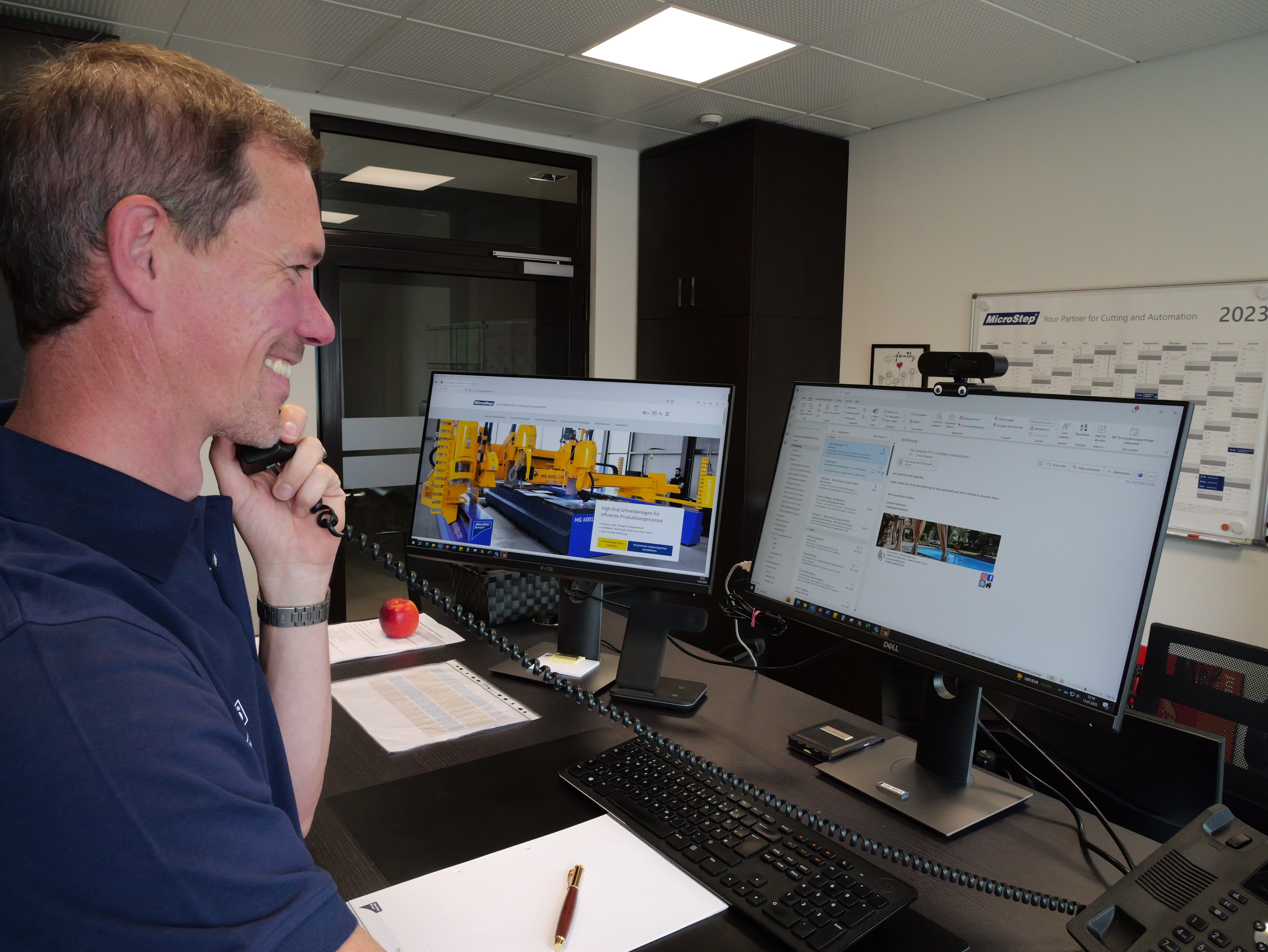 Smiling man on phone at desk with dual monitors showing email and industrial machinery website.