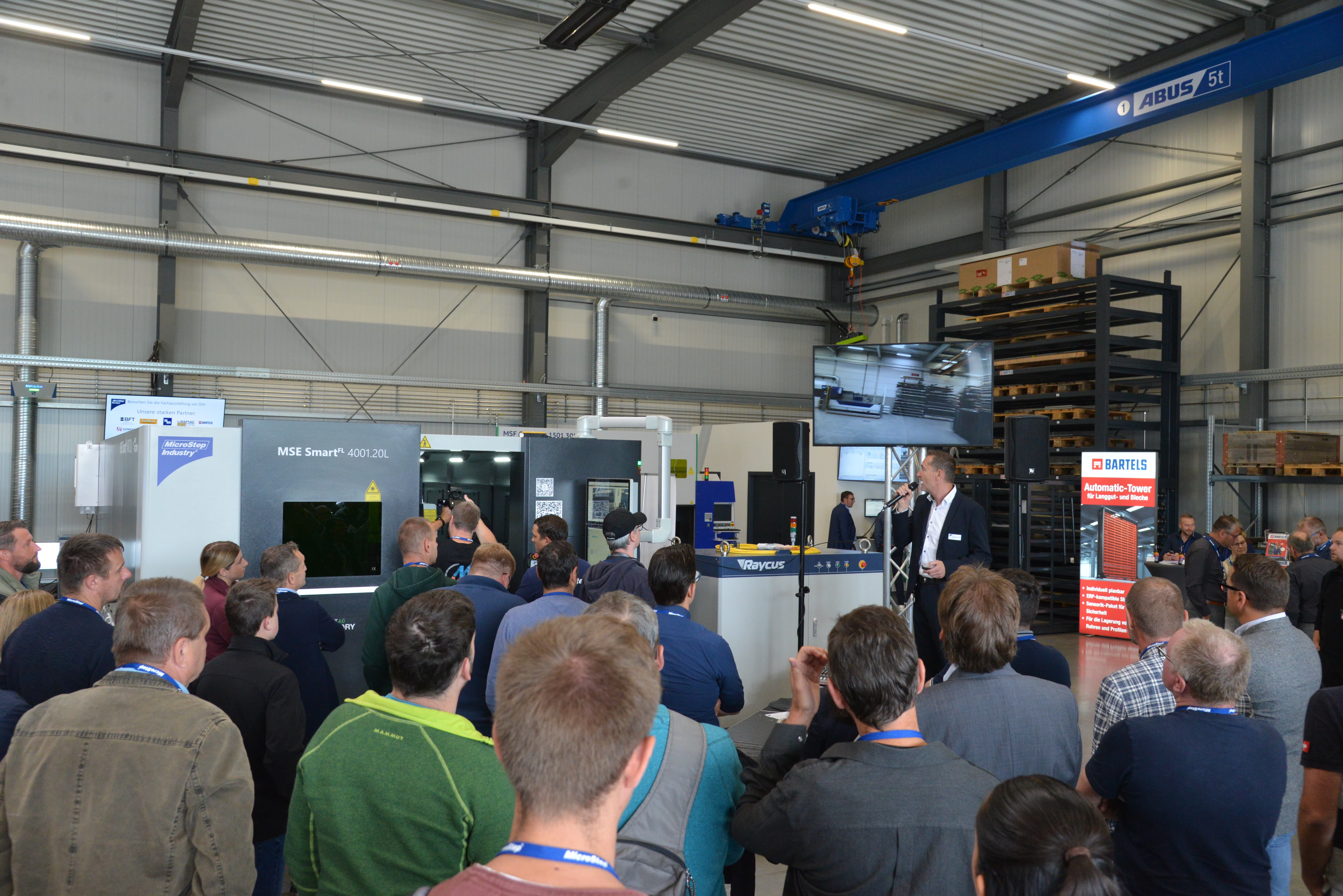 Audience listens to a man presenting industrial machinery in a large workshop or factory setting.