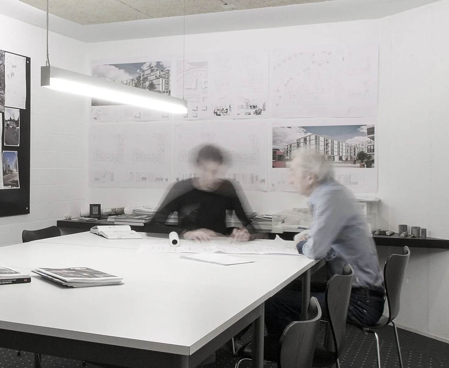 Zwei Personen sitzen an einem Tisch in einem Büro mit Architekturplänen an der Wand.