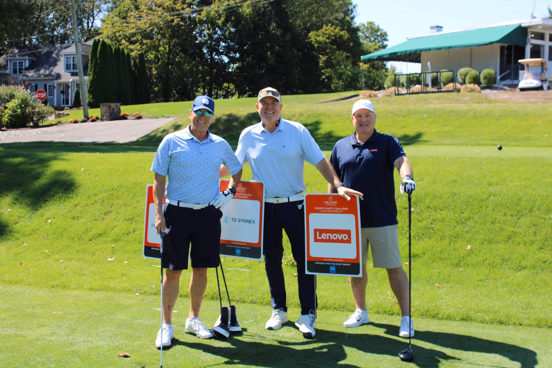Three golfers posing with Kelser Charity Challenge sponsor signs for TD Synnex and Lenovo at a golf course event
