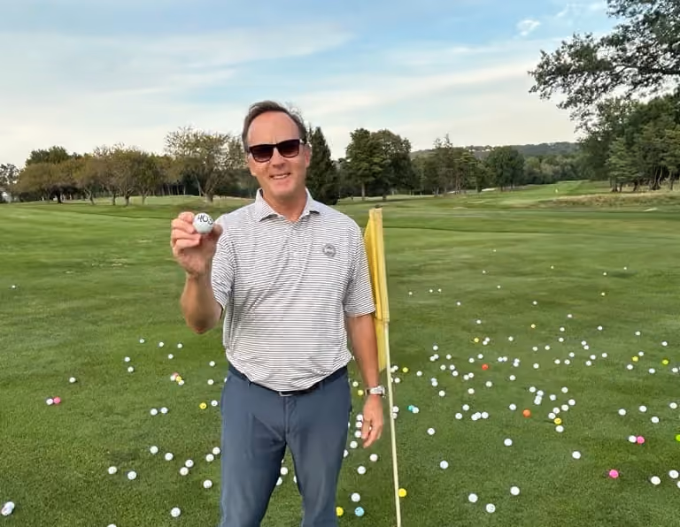Man holding golf ball on green course surrounded by scattered colorful golf balls near yellow flag, Glastonbury CT