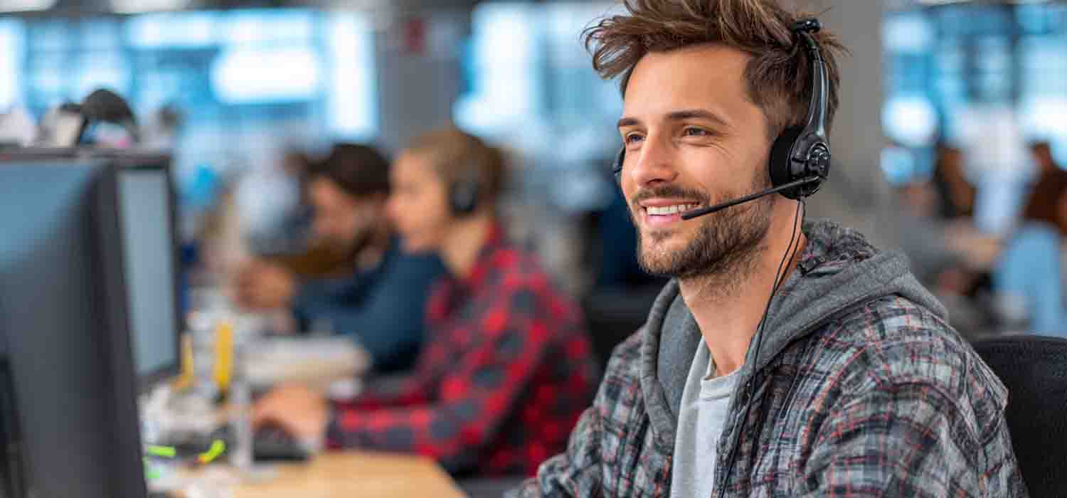 Smiling IT support technician wearing a headset working at a computer in a busy managed services help desk office