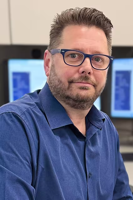 Man in blue shirt and glasses seated in front of dual computer monitors in an IT office environment