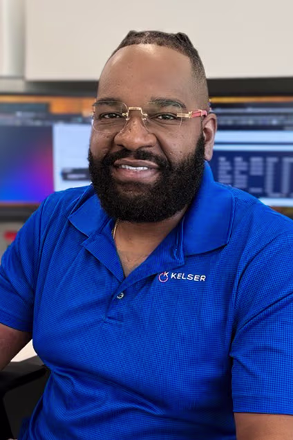 Kelser Corporation IT professional smiling at camera in branded blue polo shirt, computer monitors visible in background