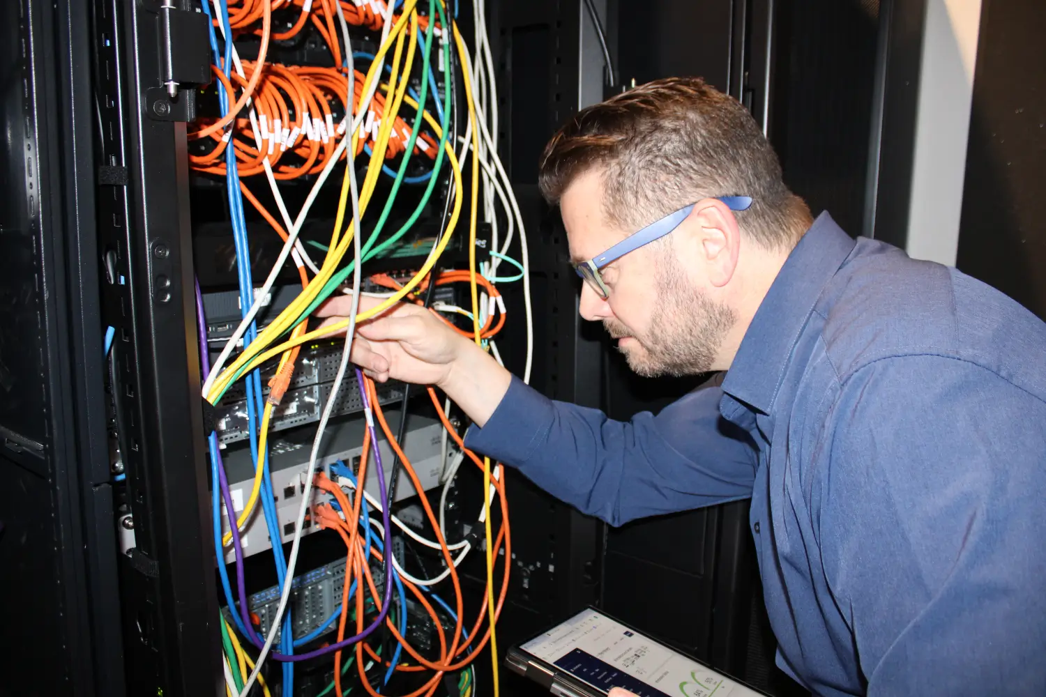 IT technician managing network cables and server rack equipment while referencing a tablet in a data center