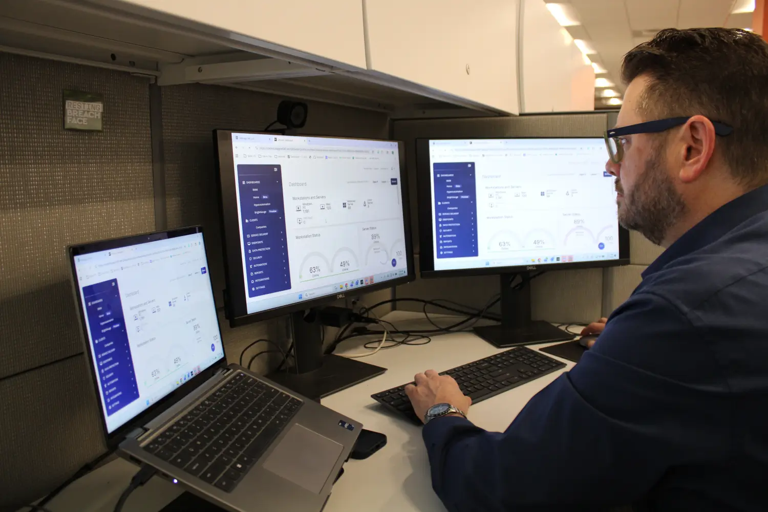 IT technician monitoring managed IT dashboards across three screens displaying server and workstation status metrics
