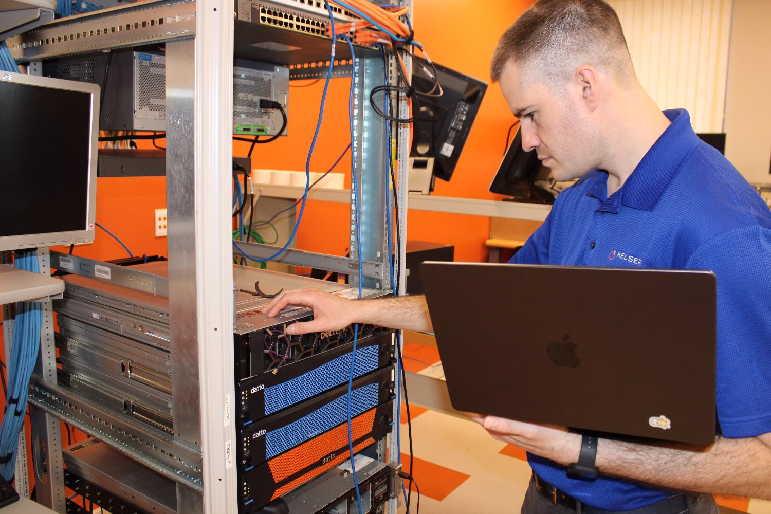 Kelser IT technician configuring rack-mounted servers and Datto backup appliances while holding a MacBook laptop
