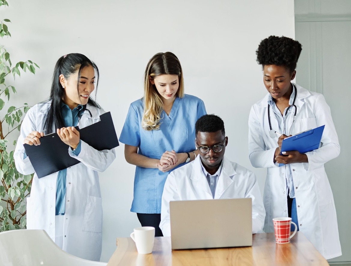 Four medical professionals collaborating around a laptop at a wooden table with mugs nearby.