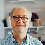 Smiling elderly man with glasses and white beard in a light blue shirt.