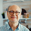 Smiling elderly man with glasses and white beard in a light blue shirt.