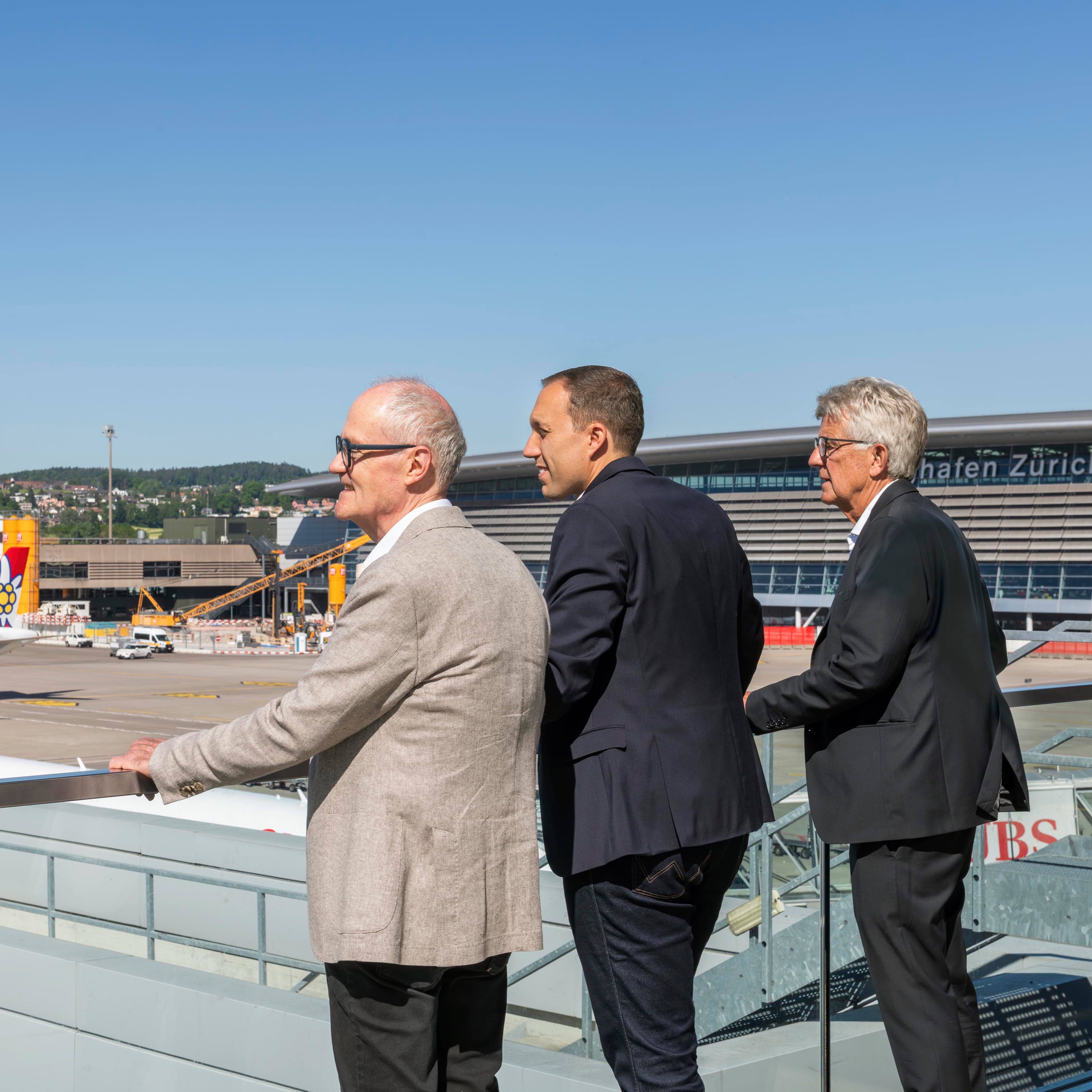 Three men in suits overlooking Zurich airport runway and terminal on a sunny day.