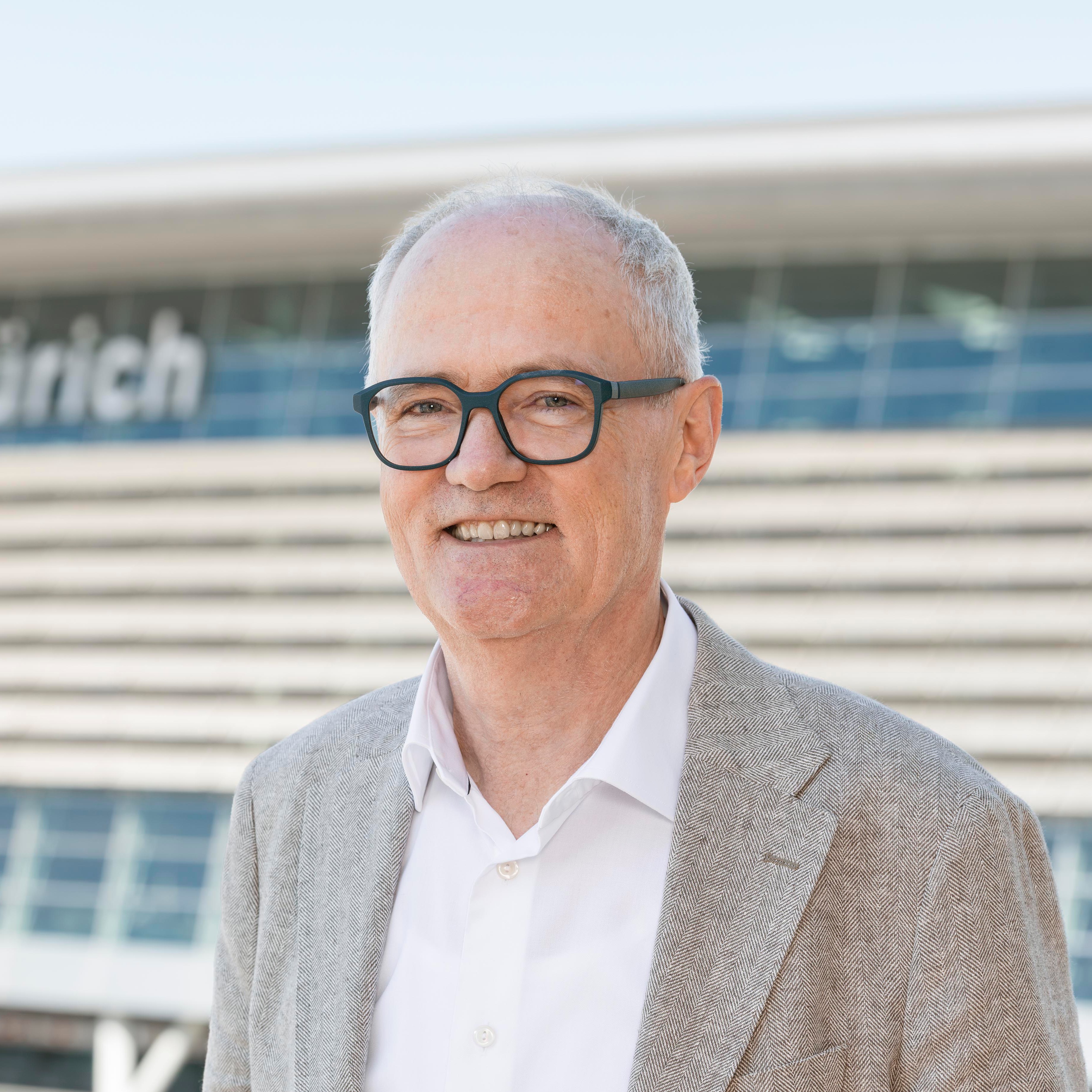 Smiling older man with glasses wearing a gray blazer and white shirt in front of a modern building.