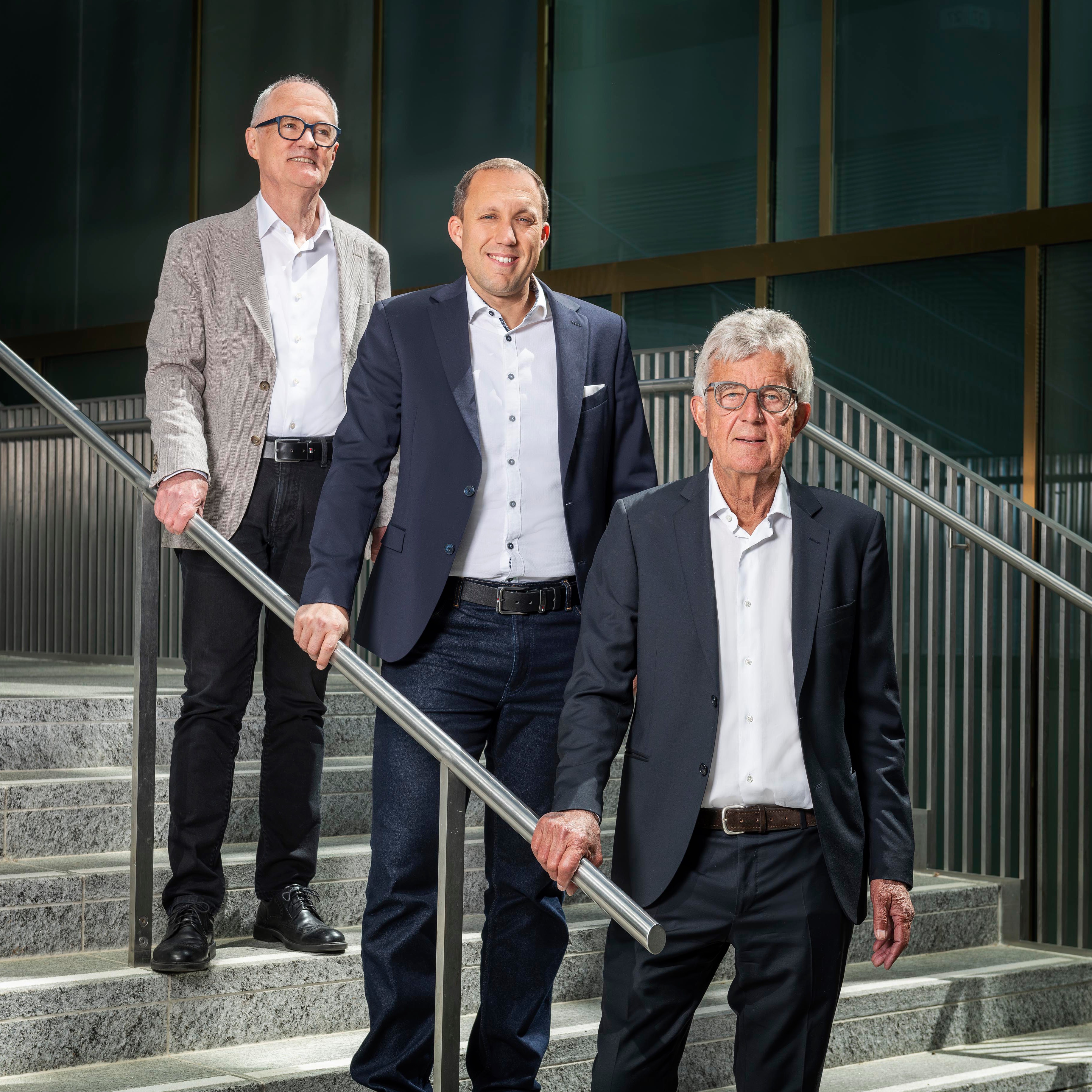 Three men in business attire standing on stairs with handrails inside a modern building.