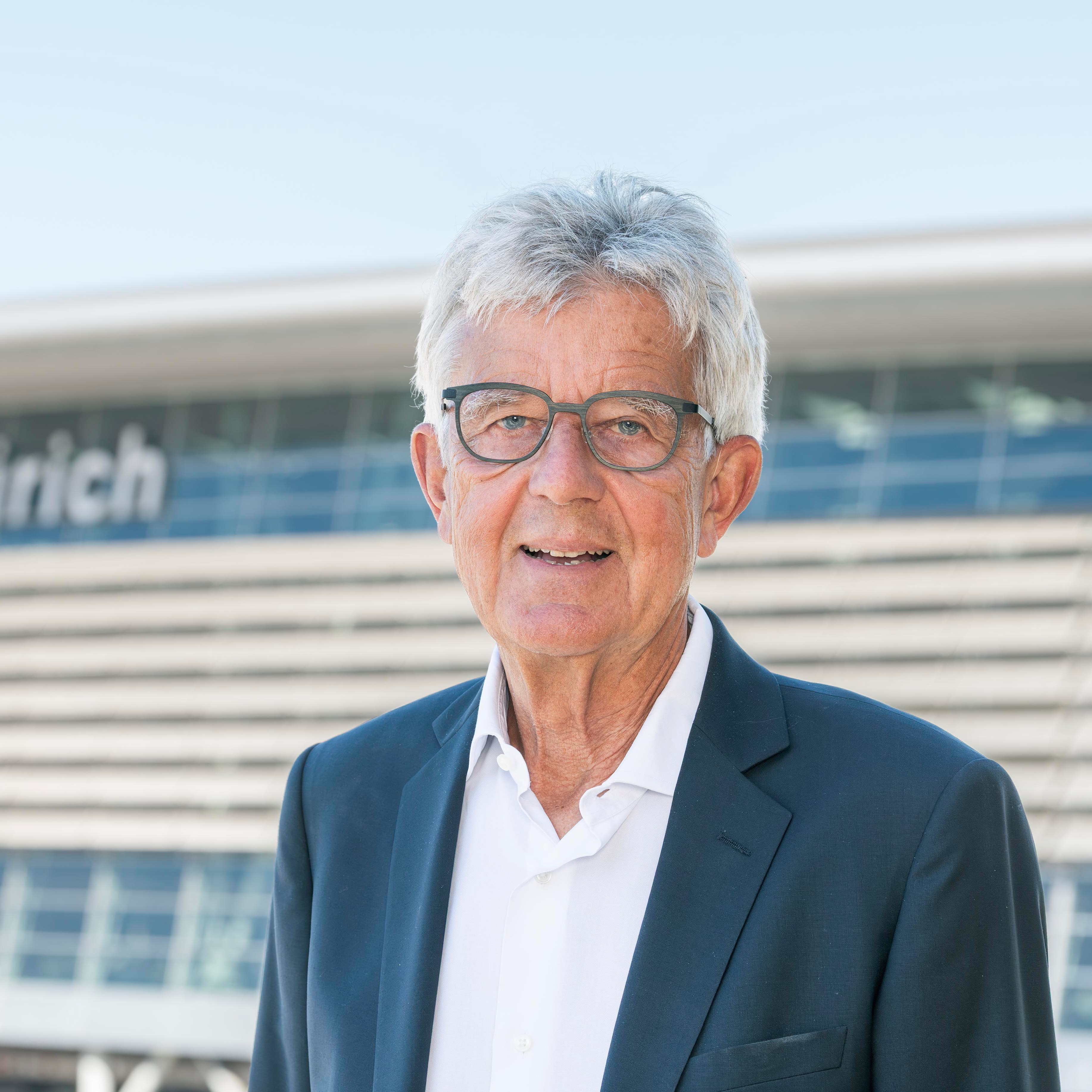 Older man with gray hair and glasses wearing a dark blazer and white shirt outdoors.