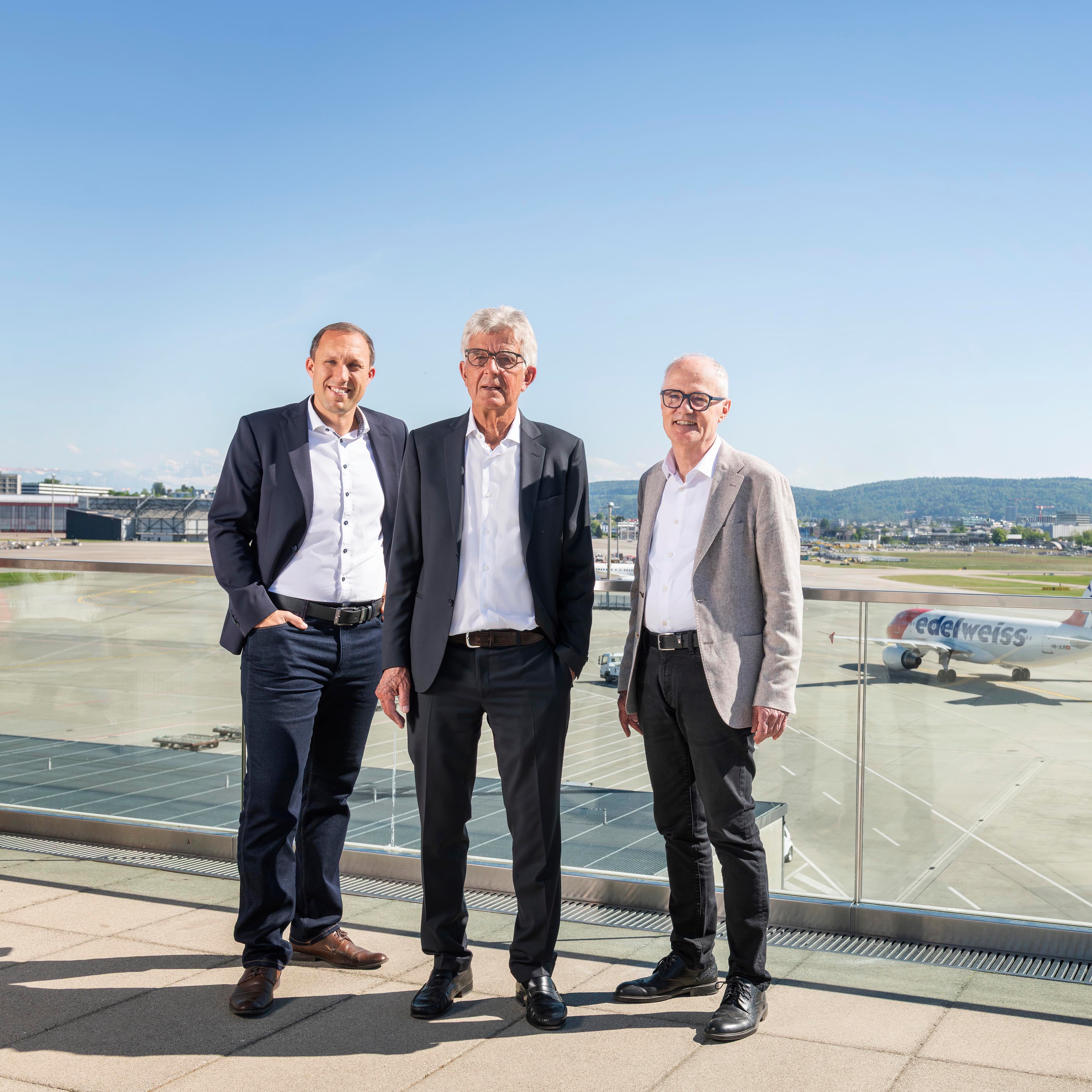 Three men in business attire standing on an airport terrace with an Edelweiss plane in the background.