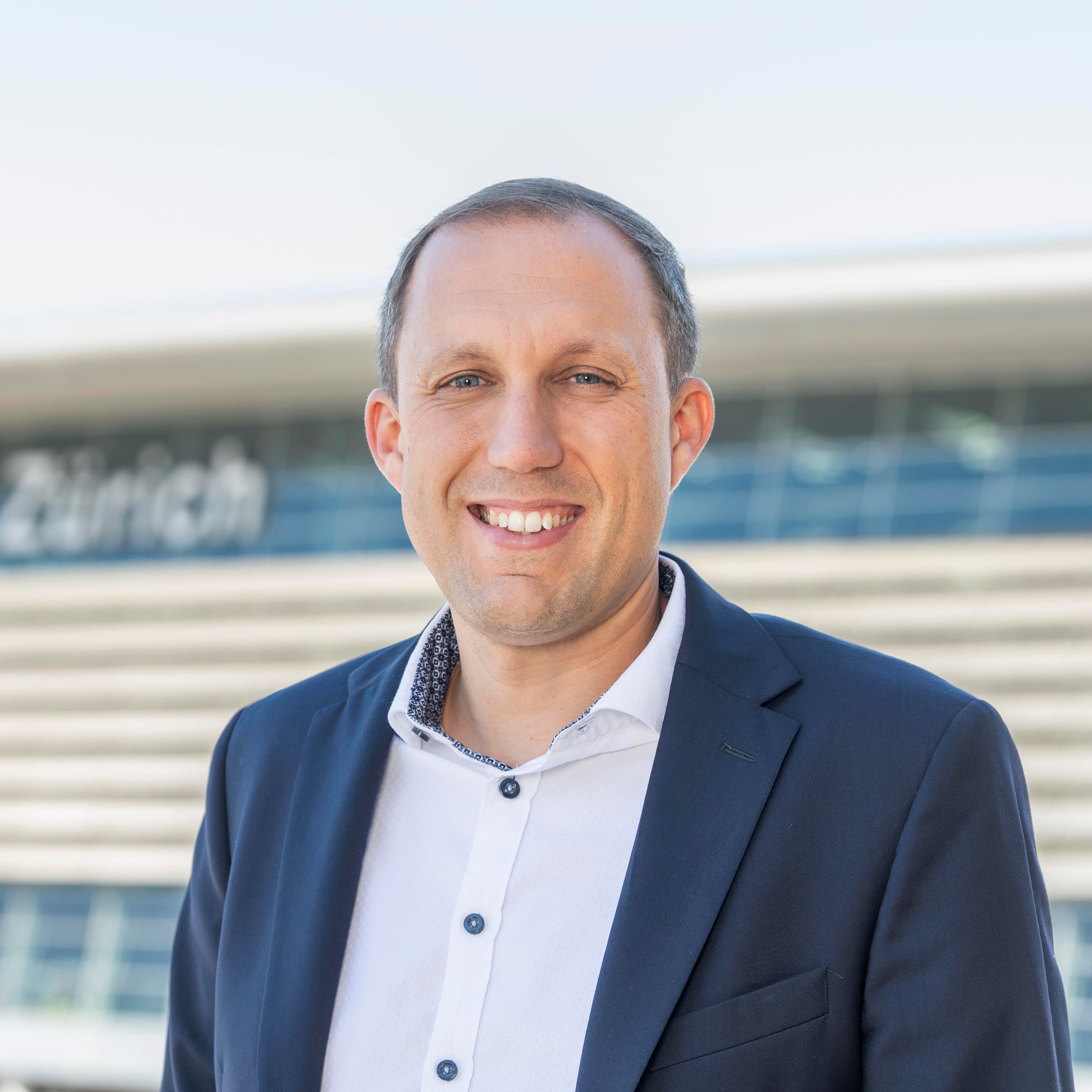 Smiling man in a navy blazer and white shirt standing outdoors with a modern building background.