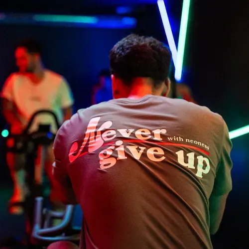 Un homme de dos portant un t-shirt avec l'inscription 'Never give up' lors d'une séance de vélo d'appartement dans une pièce sombre aux lumières néon.