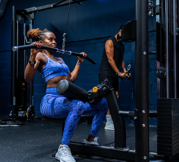 Femme sportive en tenue bleue effectuant un exercice de tirage vertical assise dans une salle de gym, homme en arrière-plan utilisant un autre appareil de musculation.