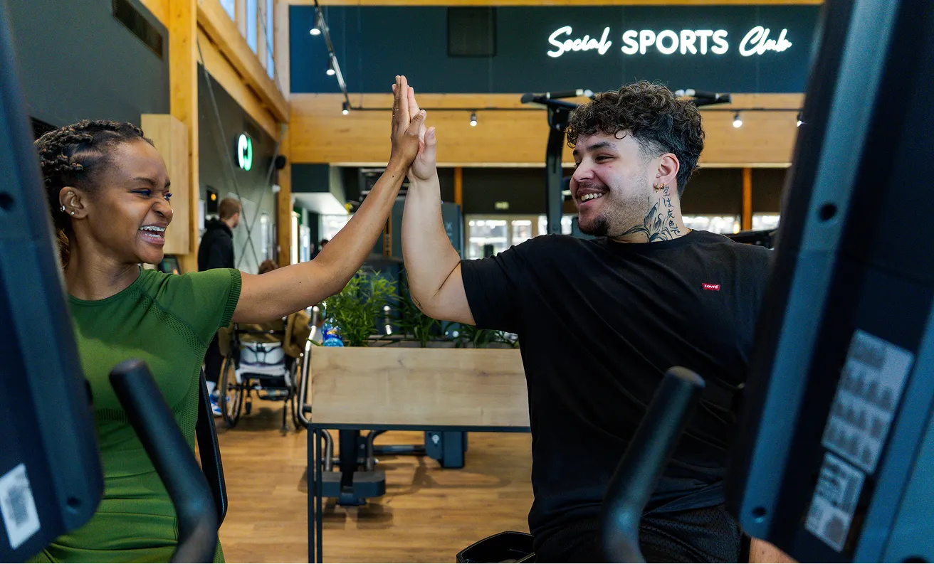 Deux personnes souriantes donnant un high five dans une salle de sport moderne équipée.