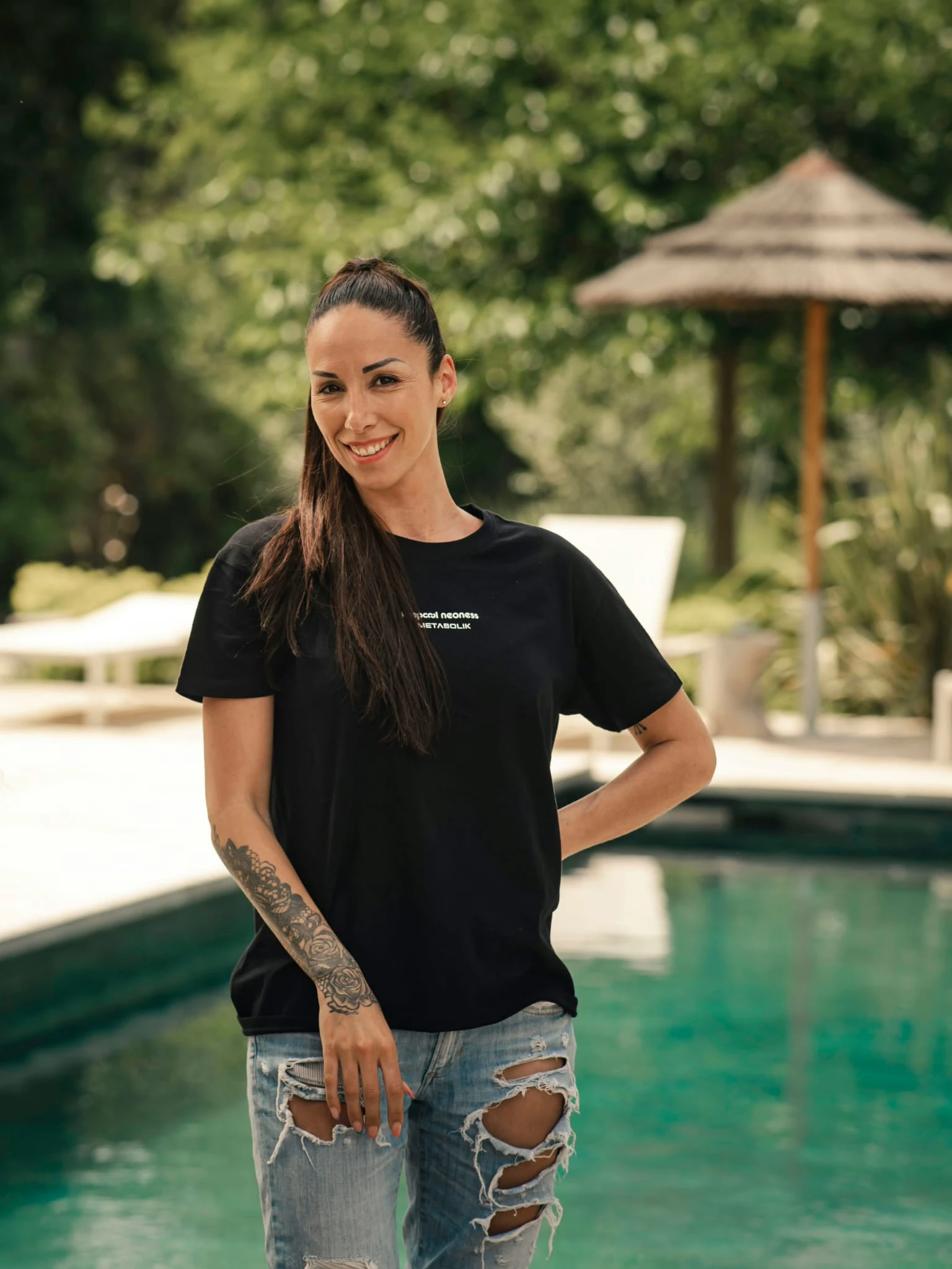 Femme souriante près piscine, t-shirt Keepcool, jeans déchirés, tatouage, cadre extérieur salle sport.