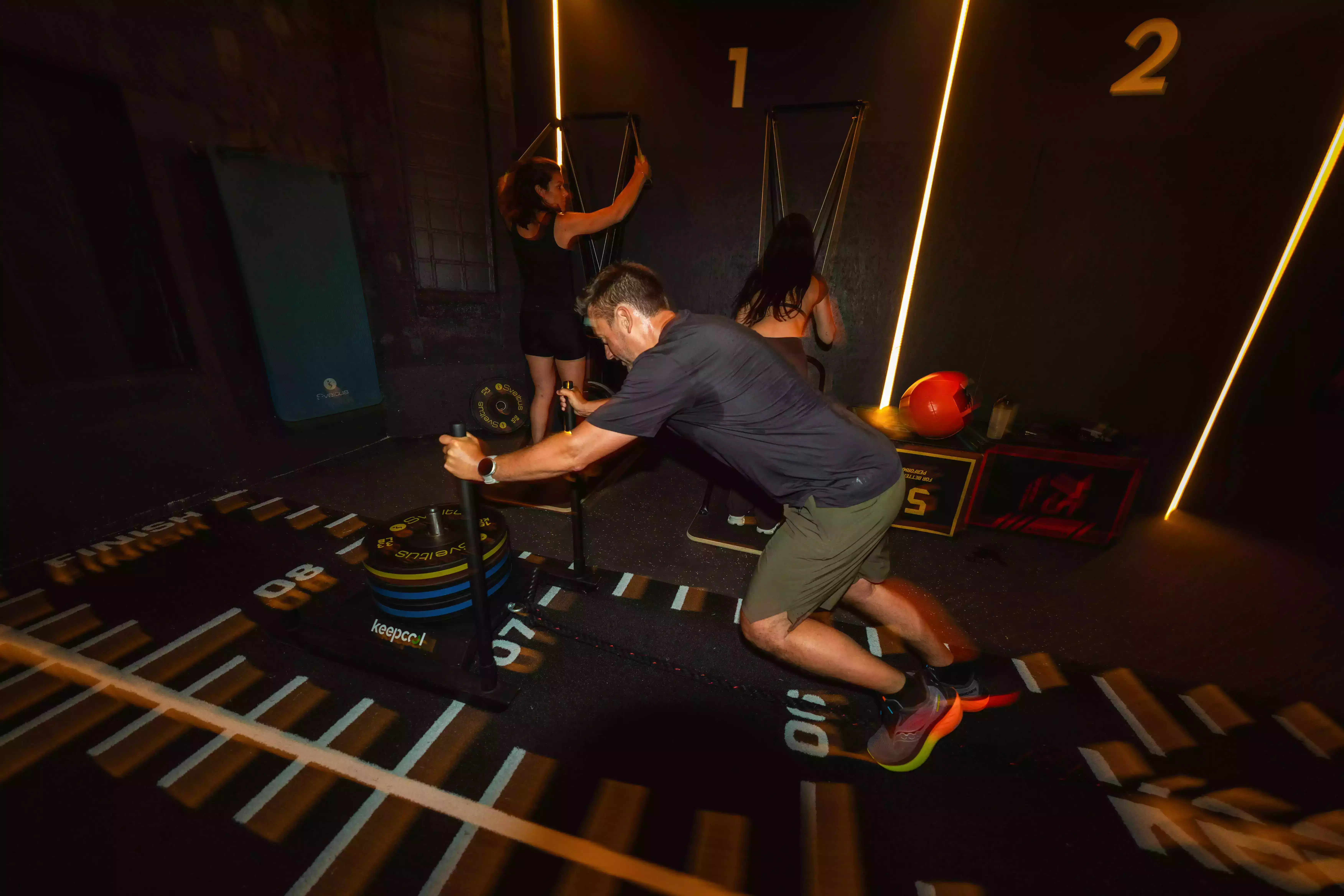 Un homme pousse un traîneau lesté pendant que deux femmes utilisent une machine de musculation en salle.