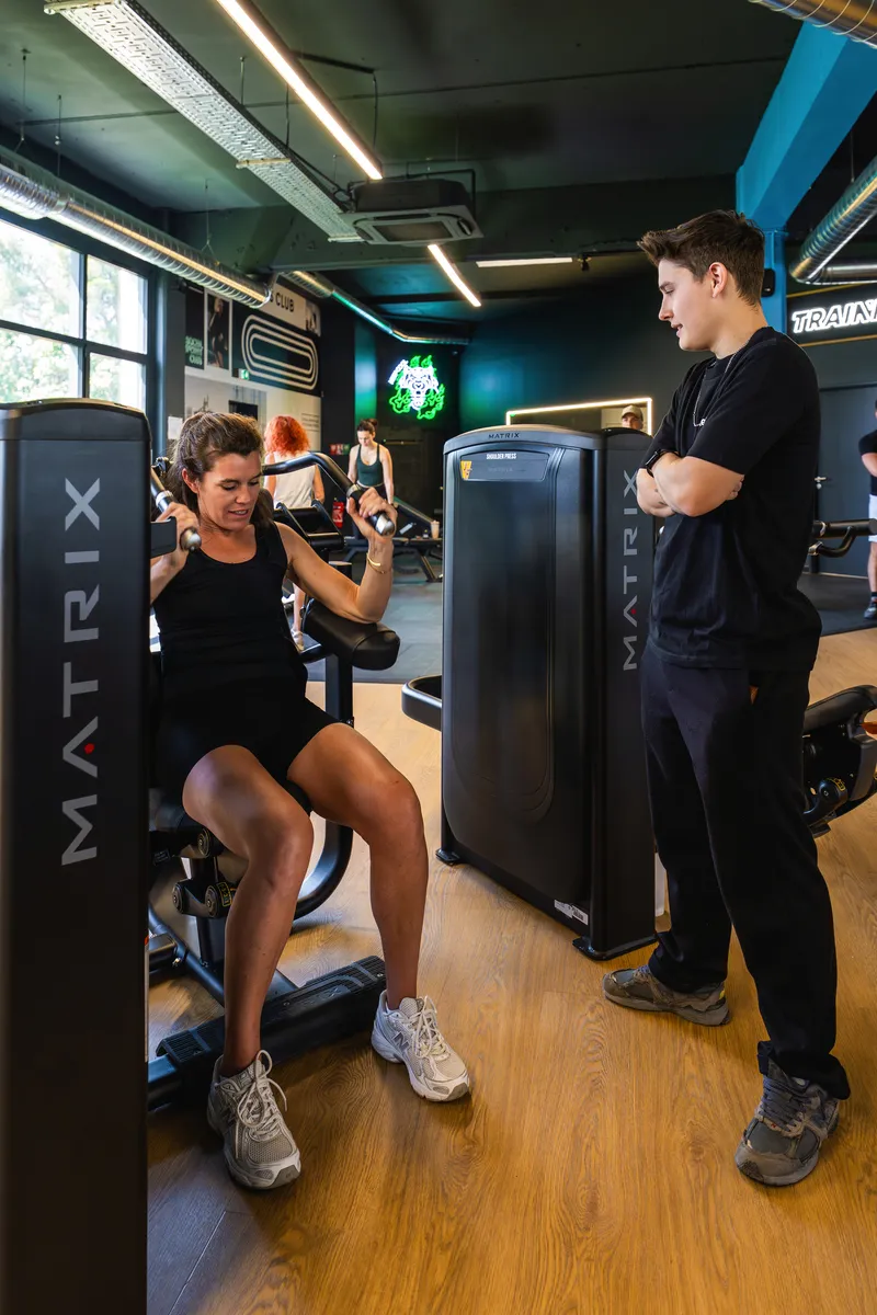 Une femme en tenue noire utilise une machine de musculation avec un coach observant dans une salle de sport moderne.