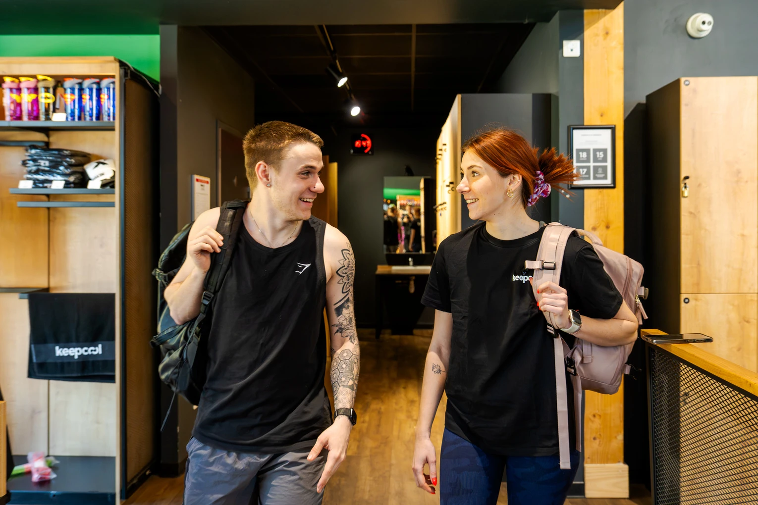 Deux personnes souriantes dans salle Keepcool, tenant boissons énergisantes, explorant espace fitness moderne avec vestiaires et équipements visibles.