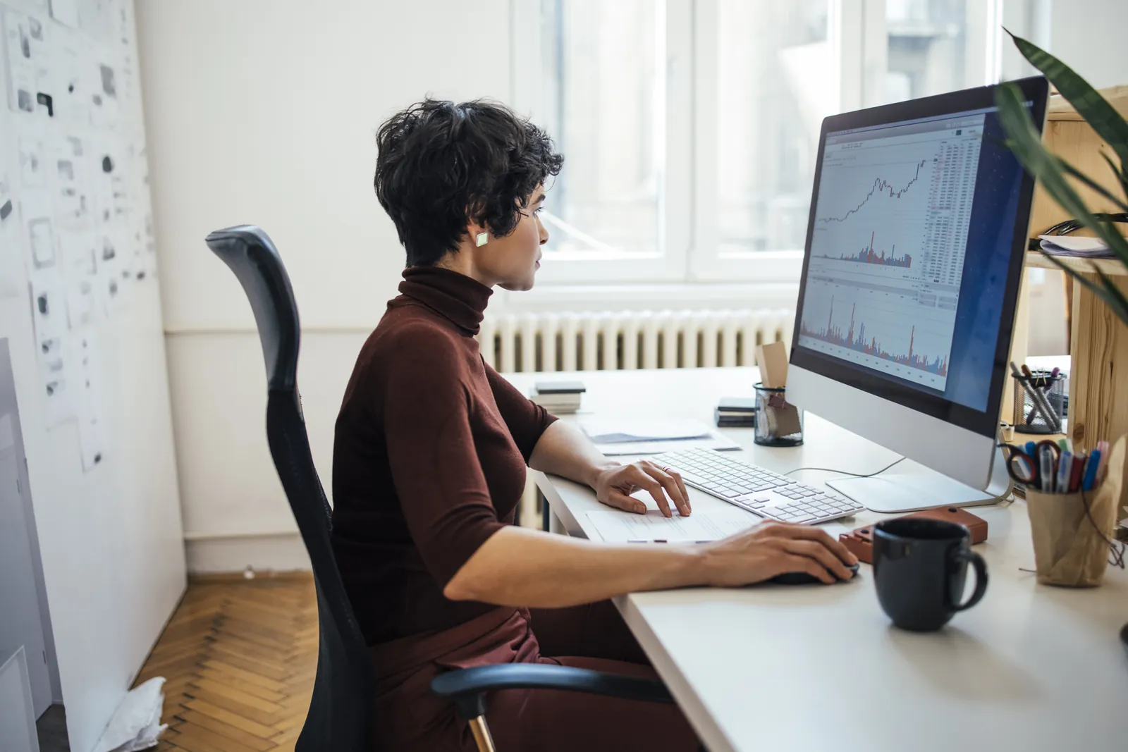 A woman at an office desk working on a desktop computer that displays financial charts and a spreadsheet