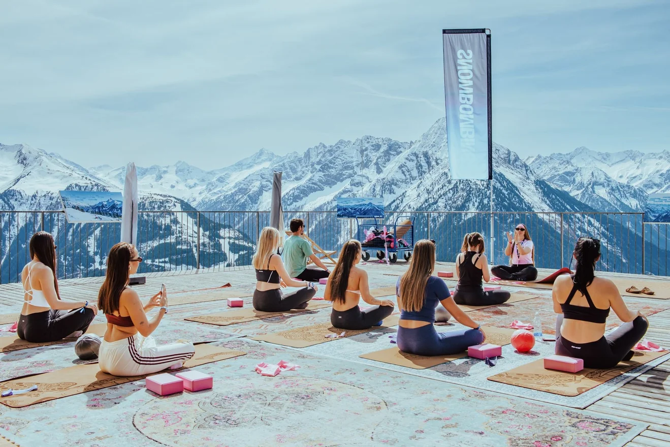 Group yoga class seated on mats on a mountaintop deck with snowy Alps in the background.