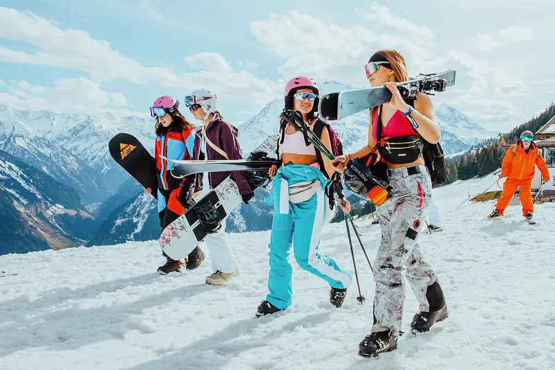 Four women carrying snowboards and skis walk on snowy mountain slope under partly cloudy sky.